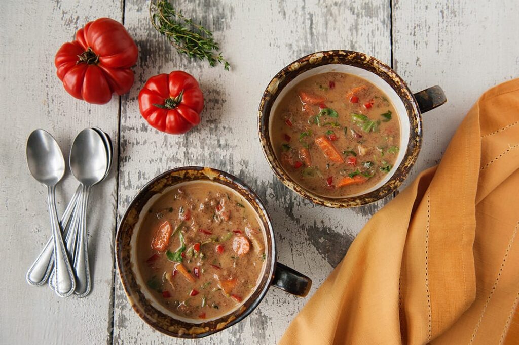 Two bowls of Gluten-Free & Vegan African Peanut Stew Recipe Sitting on table with peppers and tableware