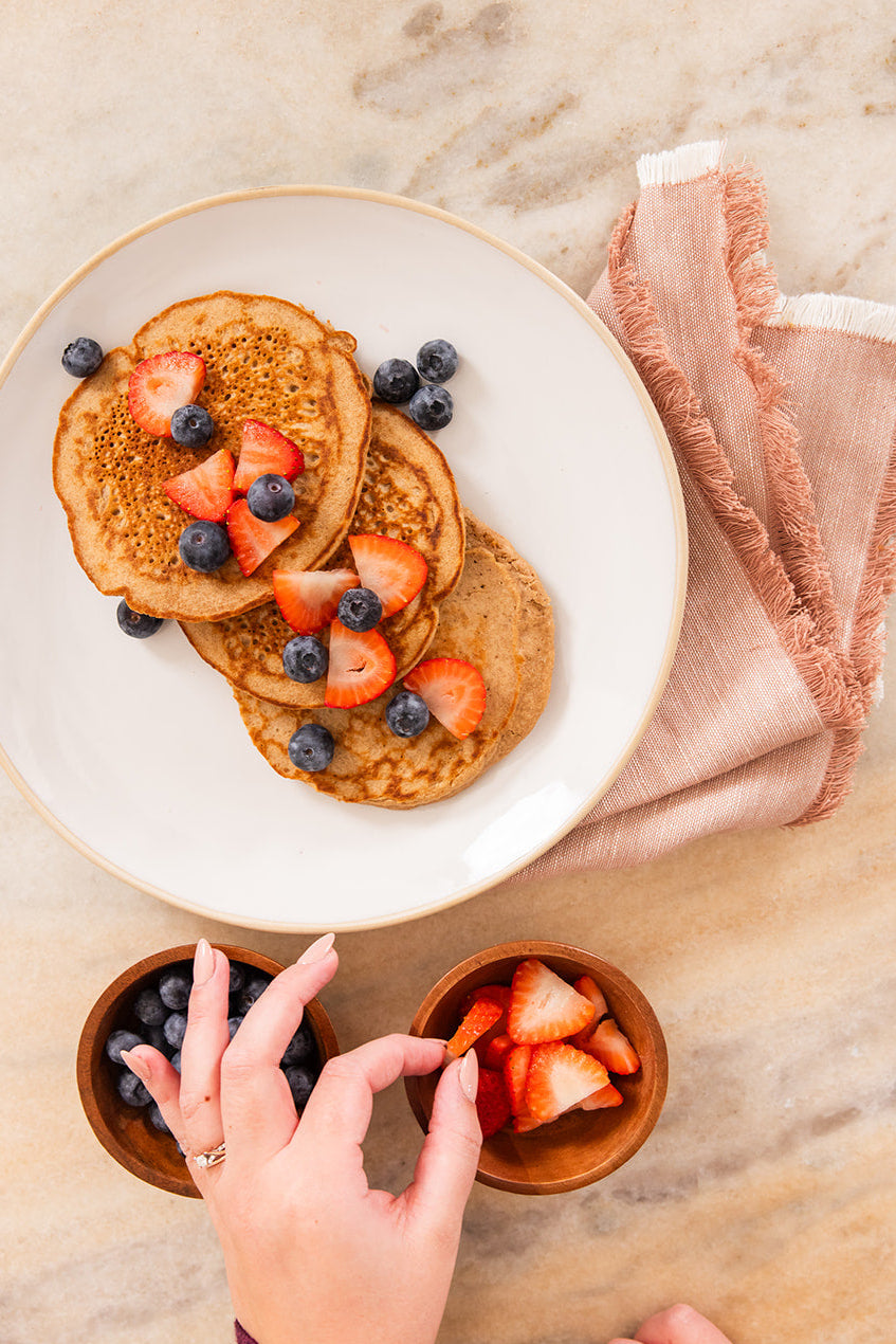 Stack of pancakes with fruit on a plate, with a hand reaching for strawberries.
