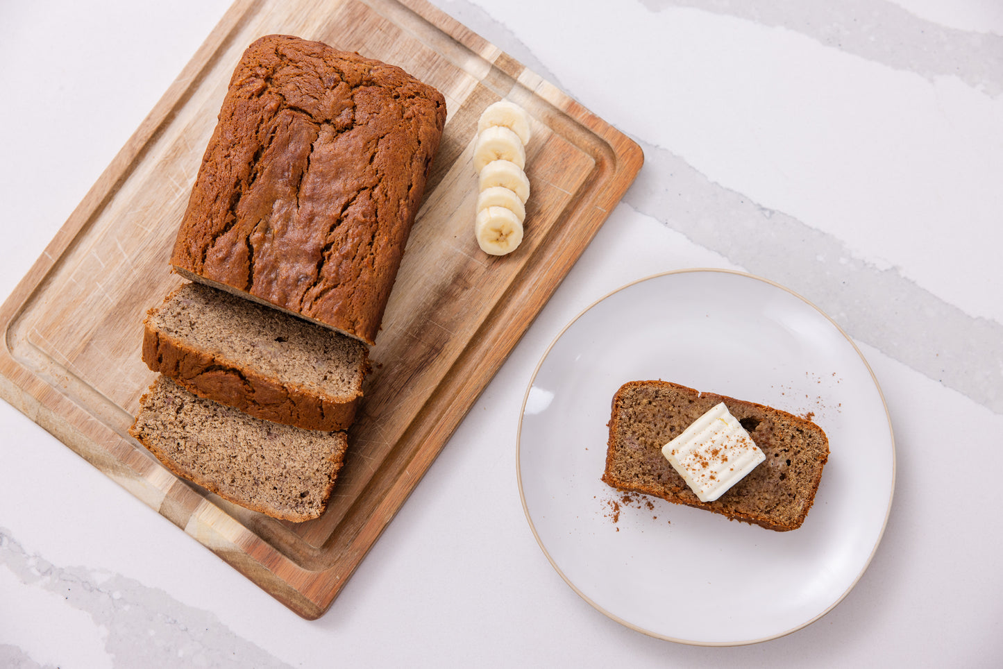 Loaf of banana bread on a wooden cutting board with slices and a slice on a plate with butter.
