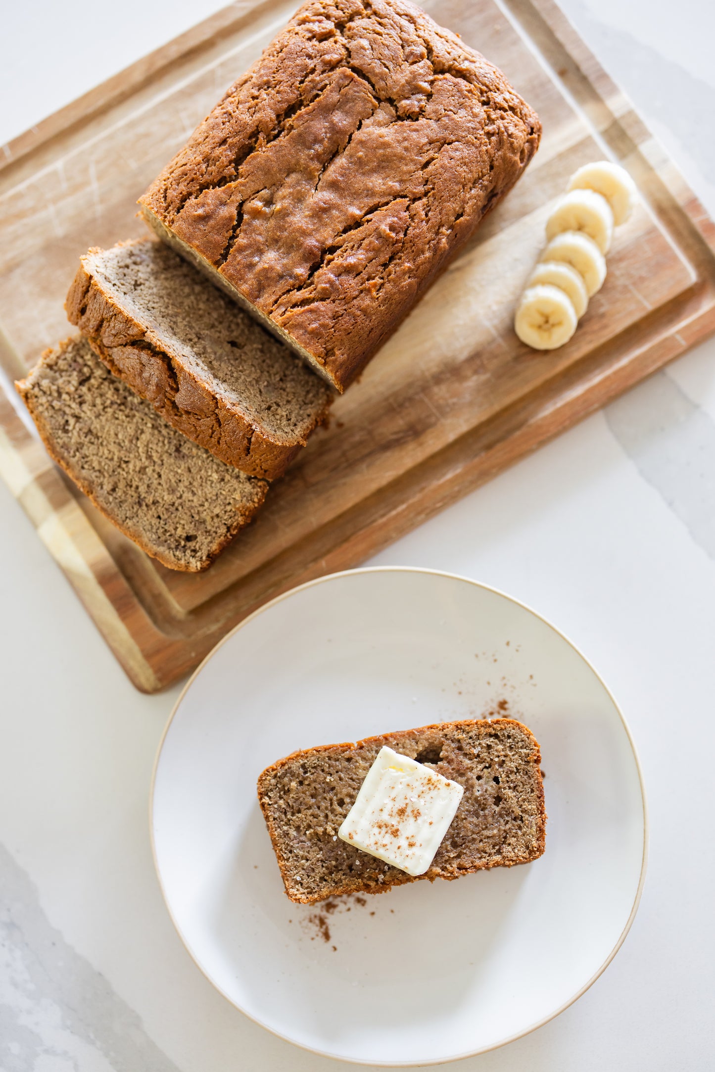 Loaf of banana bread with slices on a wooden cutting board, one slice on a plate with butter.