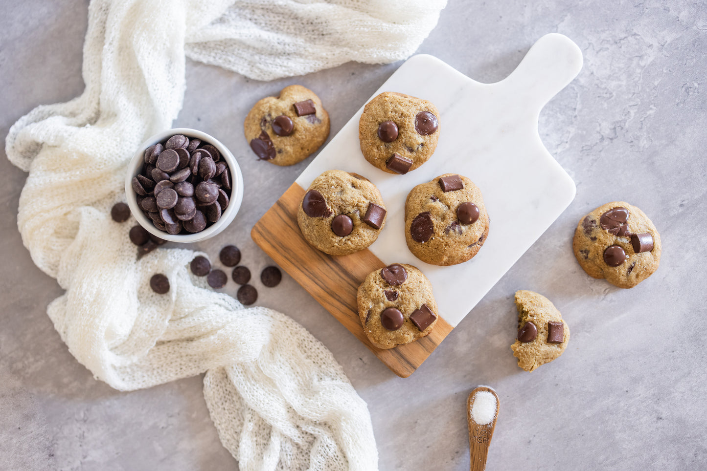 Chocolate chip cookies on a marble board with chocolate chips and a wooden spoon on a gray surface.