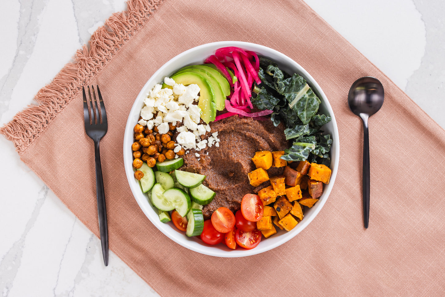 Colorful teff grain bowl with various ingredients on a pink cloth with utensils.