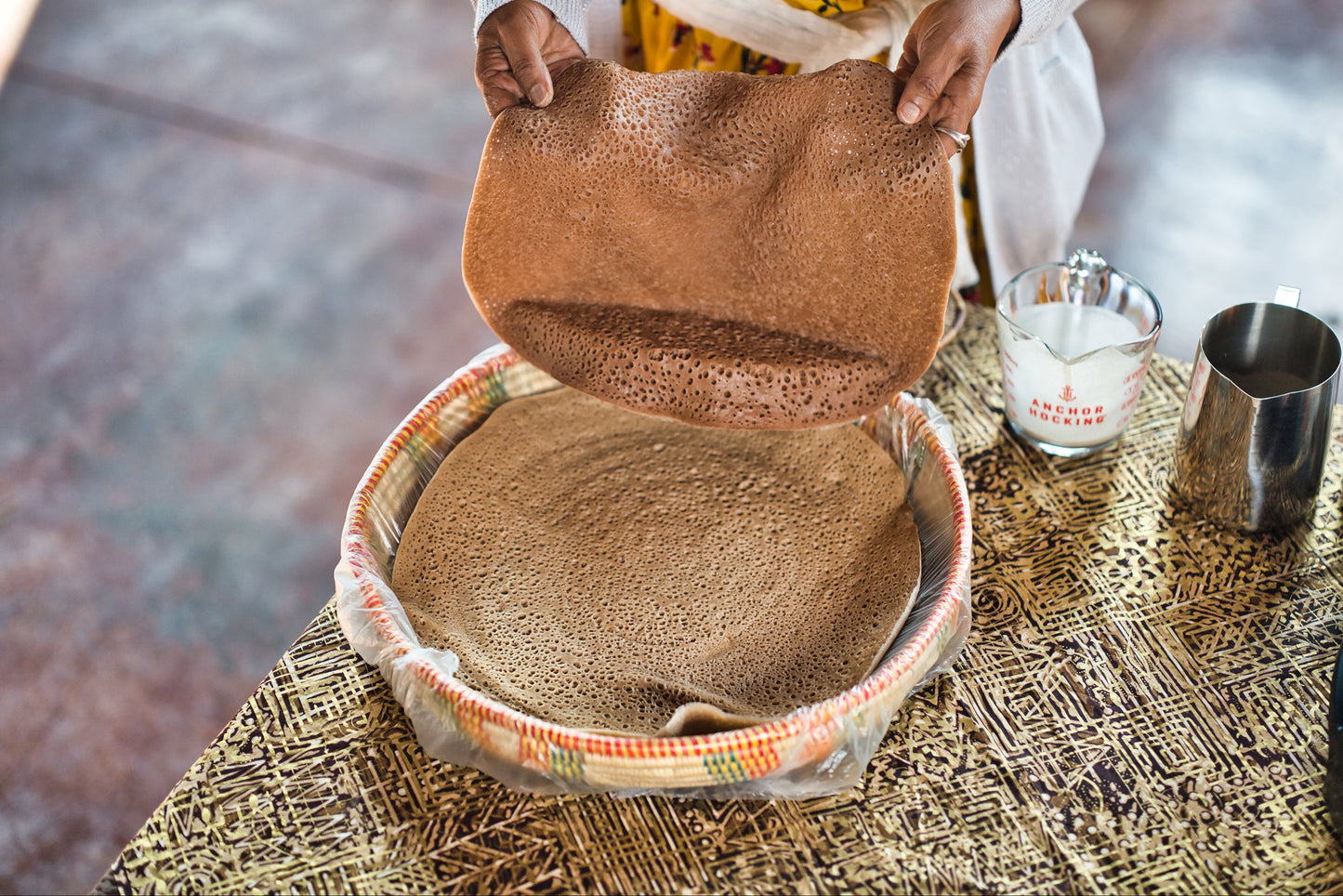 Person opening a woven basket containing a large piece of injera on a textured surface.